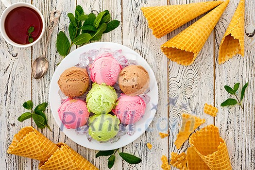 Multicolored balls of cream ice cream on an ice plate, tea and waffle cones on a wooden table.