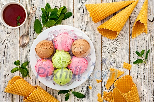 Multicolored balls of cream ice cream on an ice plate, tea and waffle cones on a wooden table. Multicolored balls of cream ice cream on an ice plate, tea and waffle cones on a wooden table.