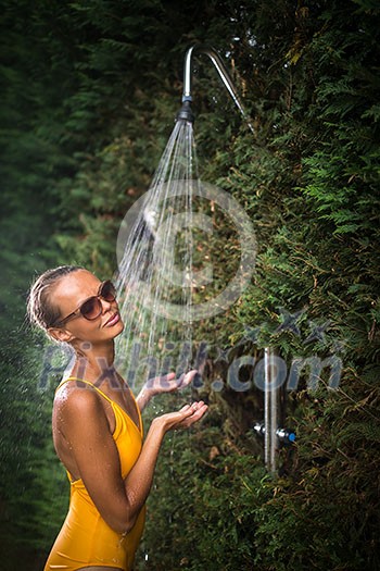 Beautiful young woman taking a shower outdoors. Attractive young woman in yellow one-piece enjoys shower on hot sunny day after a swim. Heat and refreshment concept.