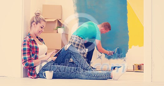 Happy couple doing home renovations, the man is painting the room and the woman is relaxing on the floor and connecting with a tablet Happy couple doing home renovations, the man is painting the room and the woman is relaxing on the floor and connecting with a tablet