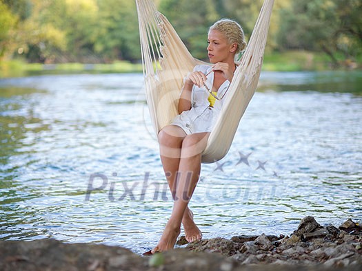 Young blonde woman resting on hammock while enjoying nature on the river bank