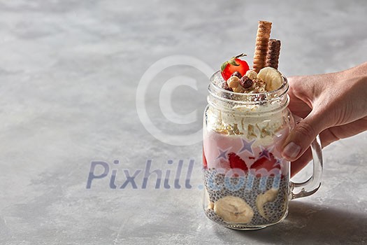 A glass with a handle in a woman's hand with a healthy breakfast of yogurt, strawberry, chia pudding with a banana, decorated with biscuits and whipped cream on a gray concrete background. Copy space
