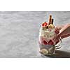 A glass with a handle in a woman's hand with a healthy breakfast of yogurt, strawberry, chia pudding with a banana, decorated with biscuits and whipped cream on a gray concrete background. Copy space