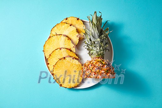 Yellow rings of sliced pineapple with a green top on a white round plate isolated on a blue background with space under the text, top view