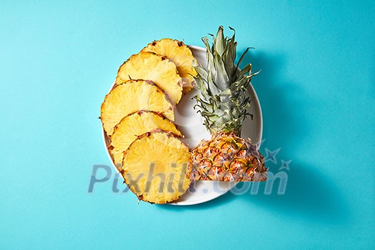 Yellow rings of sliced pineapple with a green top on a white round plate isolated on a blue background with space under the text, top view Yellow rings of sliced pineapple with a green top on a white round plate isolated on a blue background with space under the text, top view