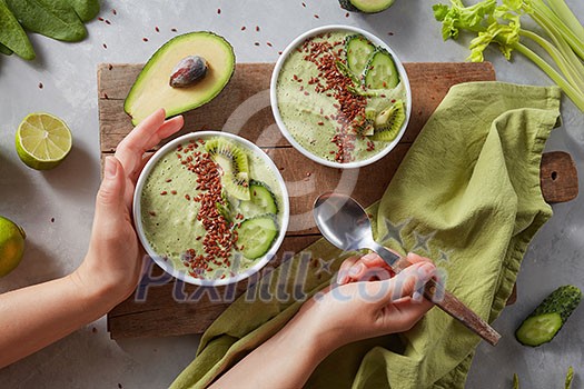 Breakfast detox green smoothie bowl with fresh kiwi, flaxseeds, cucumber and avocado in the female hands on a wooden board with a green napkin and celery sprigs, top view. Super Food Breakfast detox green smoothie bowl with fresh kiwi, flaxseeds, cucumber and avocado in the female hands on a wooden board with a green napkin and celery sprigs, top view. Super Food