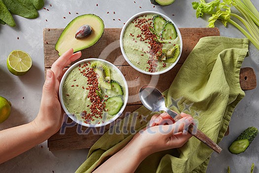 Breakfast detox green smoothie bowl with fresh kiwi, flaxseeds, cucumber and avocado in the female hands on a wooden board with a green napkin and celery sprigs, top view. Super Food