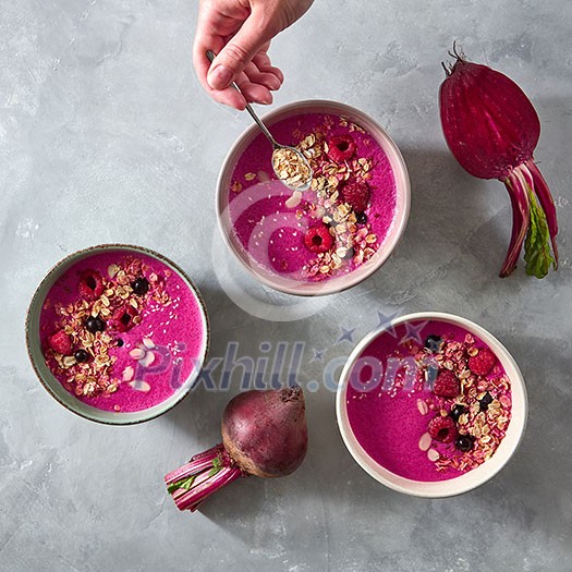 The girl's hand puts oatmeal in a plate with freshly prepared beetroot smoothies with raspberries and currants on a gray concrete background. Top view. Concept of diet and vegetarian food