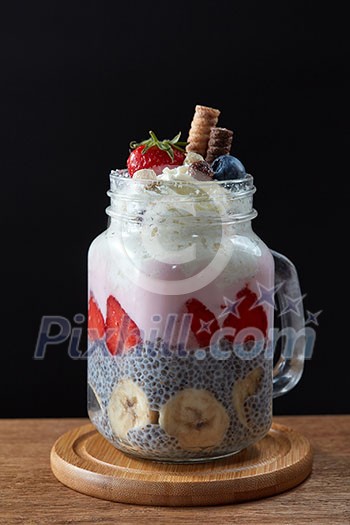 Healthy breakfast chia pudding with strawberries, banana and yogurt in glass mason jar on a dark wooden background