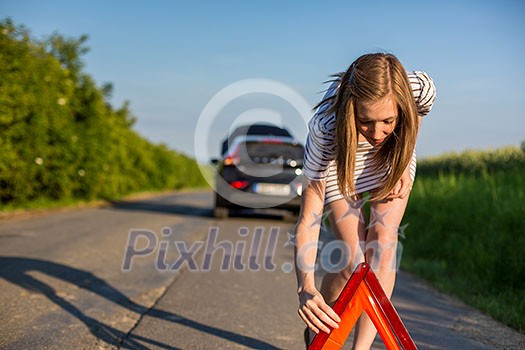 Pretty, young woman setting the safety triangle on the roadside after her car has broken down Pretty, young woman setting the safety triangle on the roadside after her car has broken down