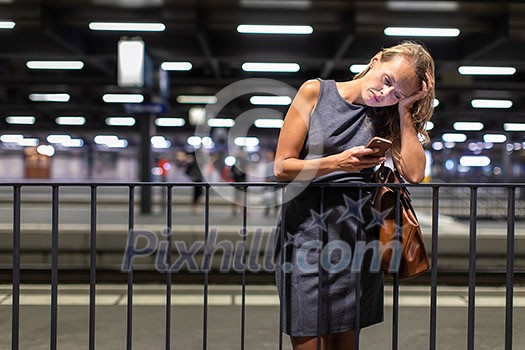Pretty, young female commuter waiting for her daily train in a modern trainstation, using her cellphone while waiting (color toned image)