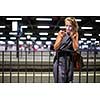 Pretty, young female commuter waiting for her daily train in a modern trainstation, using her cellphone while waiting (color toned image)