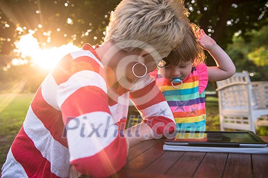 Happy mother and her little daughter enjoying free time using tablet computer while relaxing on holiday home garden during sunny day Happy mother and her little daughter enjoying free time using tablet computer while relaxing on holiday home garden during sunny day