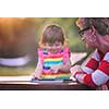 Happy mother and her little daughter enjoying free time using tablet computer while relaxing  on holiday home garden during sunny day