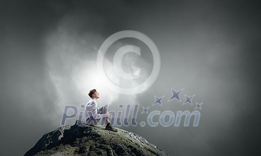 Young thoughtful businessman sitting alone on rock top Young thoughtful businessman sitting alone on rock top