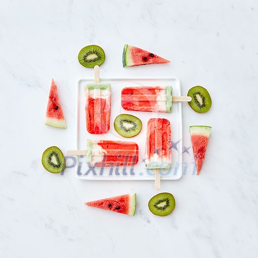 Slices of watermelon and kiwi in the form of a circle and a triangle around a plate with a healthy ice cream lolly in the shape of a square on a gray marble background with space for texas. Food pattern. Flat lay