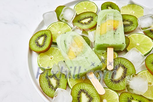 Natural organic frozen juice on a stick with a slice of mango in a bowl with pieces of lime, kiwi and ice cubes on a gray marble table with a copy of the space for text. Top view Natural organic frozen juice on a stick with a slice of mango in a bowl with pieces of lime, kiwi and ice cubes on a gray marble table with a copy of the space for text. Top view