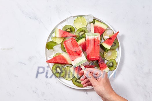 In the female hand, a healthy berry icy popsicle against the background of a gray marble table with a plate with slices of watermelon, kiwi, lime and ice cubes. Space for text. Flat lay