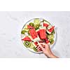In the female hand, a healthy berry icy popsicle against the background of a gray marble table with a plate with slices of watermelon, kiwi, lime and ice cubes. Space for text. Flat lay