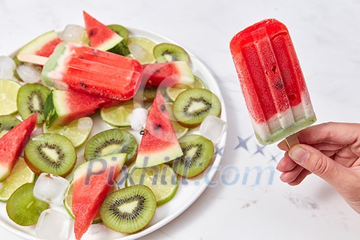 A woman's hand holds berry ice cream lolly, on a white background a plate of ice cream, pieces of kiwi, watermelon and ice cubes. Copy space for text A woman's hand holds berry ice cream lolly, on a white background a plate of ice cream, pieces of kiwi, watermelon and ice cubes. Copy space for text