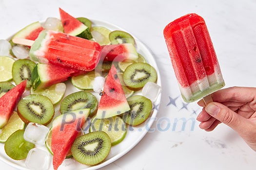 A woman's hand holds berry ice cream lolly, on a white background a plate of ice cream, pieces of kiwi, watermelon and ice cubes. Copy space for text