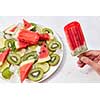 A woman's hand holds berry ice cream lolly, on a white background a plate of ice cream, pieces of kiwi, watermelon and ice cubes. Copy space for text