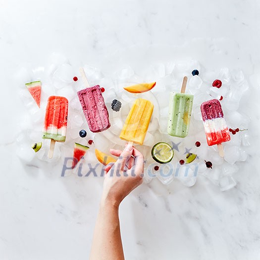 A set of different frozen berry smoothies lolly on a stick with years and fruits. The girl's hand takes a smoothie on a stick from ice cubes. Flat lay