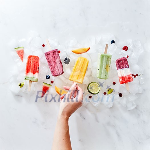 A set of different frozen berry smoothies lolly on a stick with years and fruits. The girl's hand takes a smoothie on a stick from ice cubes. Flat lay A set of different frozen berry smoothies lolly on a stick with years and fruits. The girl's hand takes a smoothie on a stick from ice cubes. Flat lay