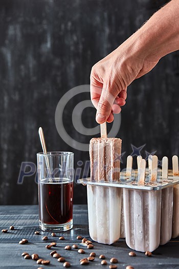 Coffee ice lolly. A woman's hand takes ice cream with plastic molds, a glass of coffee and coffee beans on a black wooden table. Copy of space