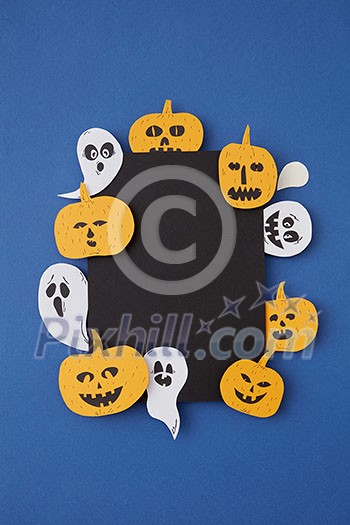 Greeting decorative card handcraft with paper horrible smiling spirits and ghosts and laughing pumpkin on a blue paper background, place under text. Top view.