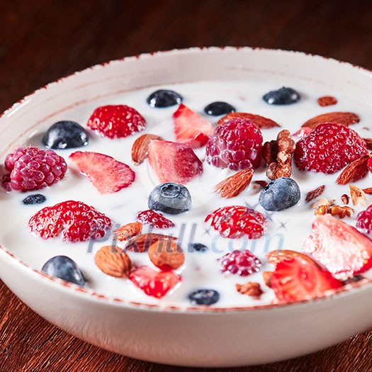 Preparing of natural breakfast with fresh organic ingredients - berries, granola, nuts, honey and pouring milk in a white bowl on a wooden table. Vegetarian detox eating