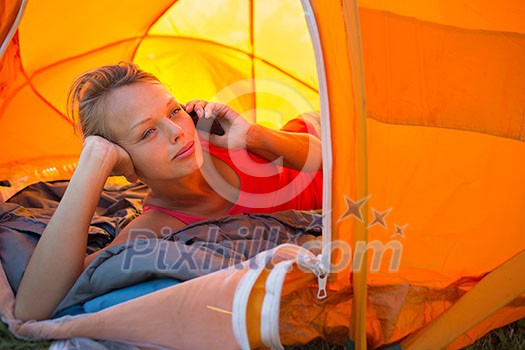 Pretty, young woman camping outdoors, lying in the tent in the morning, using her smart phone