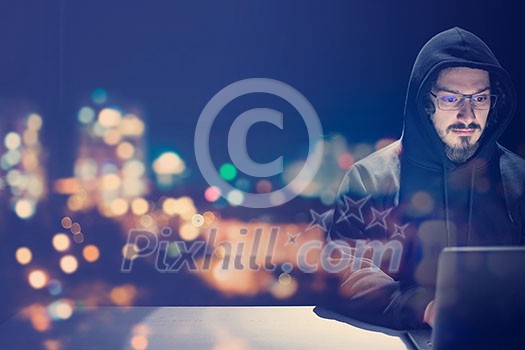 Young talented hacker using laptop computer while working in dark office with big city lights in the background at night Young talented hacker using laptop computer while working in dark office with big city lights in the background at night