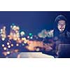 Young talented hacker using laptop computer while working in dark office with big city lights in the background at night