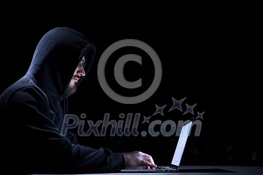 Young talented hacker using laptop computer while working in dark office with big city lights in the background at night