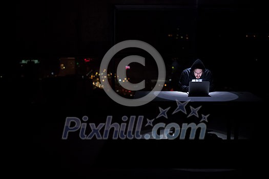 Young talented hacker using laptop computer while working in dark office with big city lights in the background at night