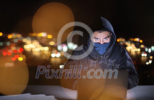 Young talented hacker using laptop computer while working in dark office with big city lights in the background at night
