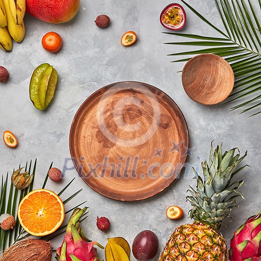 Bright composition of various exotic fruits, palm green leaves and brown wooden empty plates on a gray concrete background with copy space. Flat lay