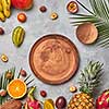 Bright composition of various exotic fruits, palm green leaves and brown wooden empty plates on a gray concrete background with copy space. Flat lay