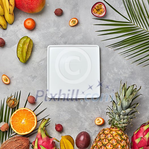 Juicy ripe tropical fruits, pineapple, papaya, mango, pineapple, green palm leaves and empty white square plate on a gray concrete background with space for text. Flat lay