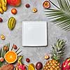 Juicy ripe tropical fruits, pineapple, papaya, mango, pineapple, green palm leaves and empty white square plate on a gray concrete background with space for text. Flat lay