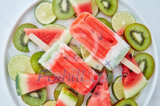 Summer dessert from frozen fruit juice lolly with ice and pieces of watermelon, kiwi and lime in a plate on a gray marble background. Flat lay