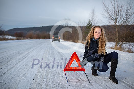 Young woman setting up a warning triangle and calling for assistance after her car broke down in the middle of nowhere on a freezing winter day