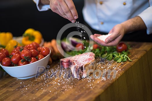 Master Chef hands putting salt on juicy slice of raw steak with vegetables around on a wooden table