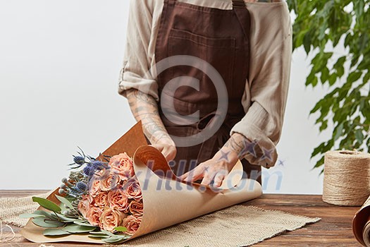 Girl's florist hands with tatoo are making bouquet with fresh flowers roses living coral color at the table with paper and rope. Process step by step. Concept floral shop.