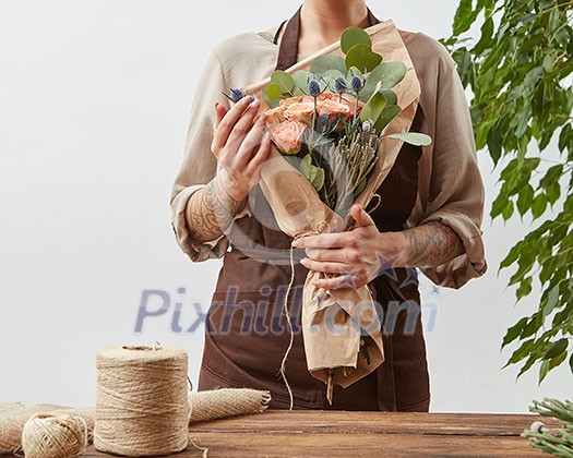 Young girl florist with tattoo hands hold bouquet with fresh flowers roses living coral color in a paper on a gray wall background. Concept floral shop and small business. Mother's Day holiday.