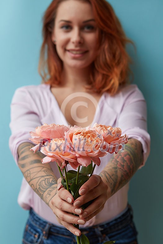 A happy red-haired woman with a tattoo holds in her hands delicate pink roses on a blue background with copy space for text. Holiday gift concept