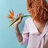 Sexy, red-haired woman with a tattoo holding an exotic strelitzia flower on a blue background with copy space. Valentine's day card