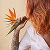 Orange strelitzia flower on the shoulder of a red-haired young girl with a tattoo around a beige background with copy space. Postcard