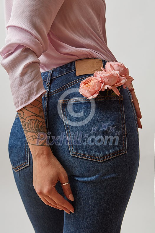 Beautiful female wearing in jeans with fresh roses of living coral color in a back pocket and pink shirt on a light gray background. Place for text. Concept of Woman's or Mother's Day.